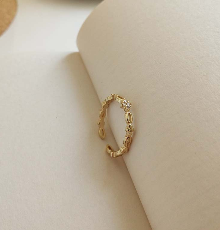 Slim gold-tone open adjustable ring with a zircon-style stone, resting on an open book