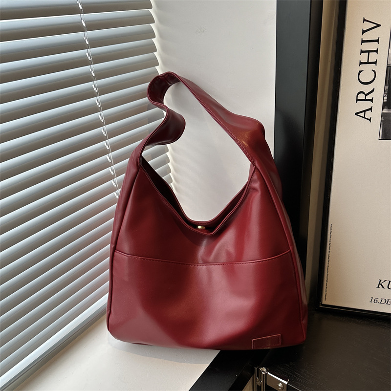 Wine red slouchy shoulder bag with a wide single strap and front seam, shown by a window with blinds
