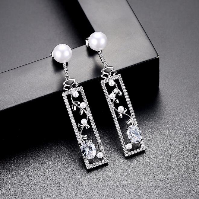 Pair of silver-tone drop earrings with zircon-style stones and pearl-style tops beside a black block