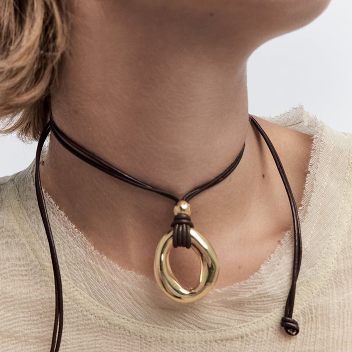 Gold-tone irregular circle pendant on a dark cord, worn with a cream top
