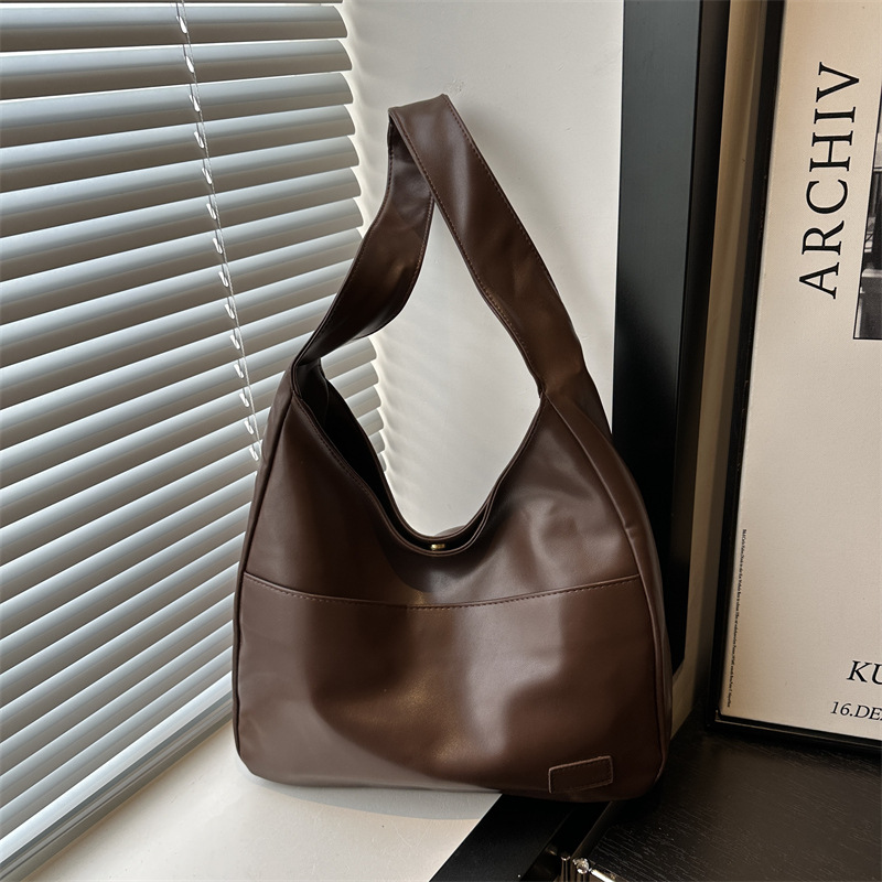 Brown slouchy shoulder bag with a wide single strap and front seam, shown by a window with blinds