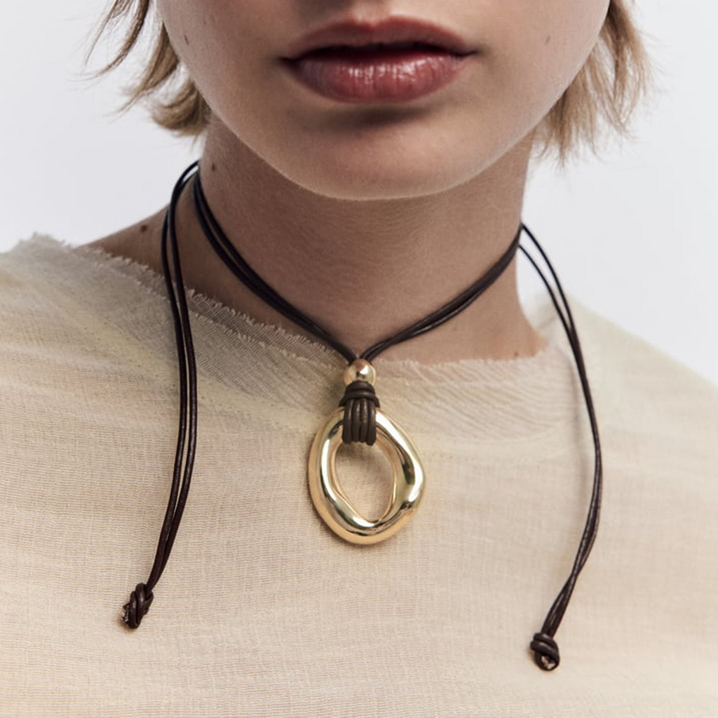 Gold-tone irregular circle pendant on a dark cord, cropped view, worn with a cream top